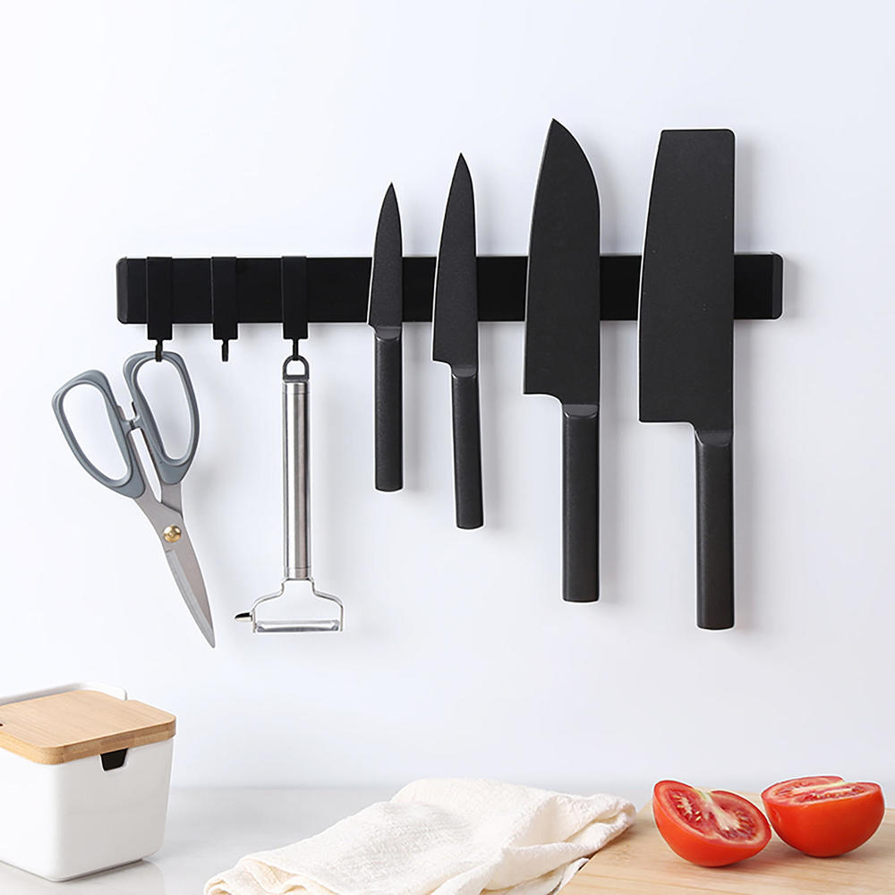 Black magnetic strip mounted on a wall holding kitchen scissors, a peeler, and four knives above a countertop with tomatoes and a storage container.