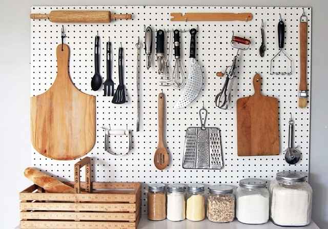 White pegboard on a wall holding various kitchen utensils, cutting boards, and graters, with jars of dry goods lined up on the counter below.